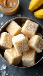 Homemade frozen peanut butter banana dog treats cubes in metal bowl with fresh bananas and peanut butter jar